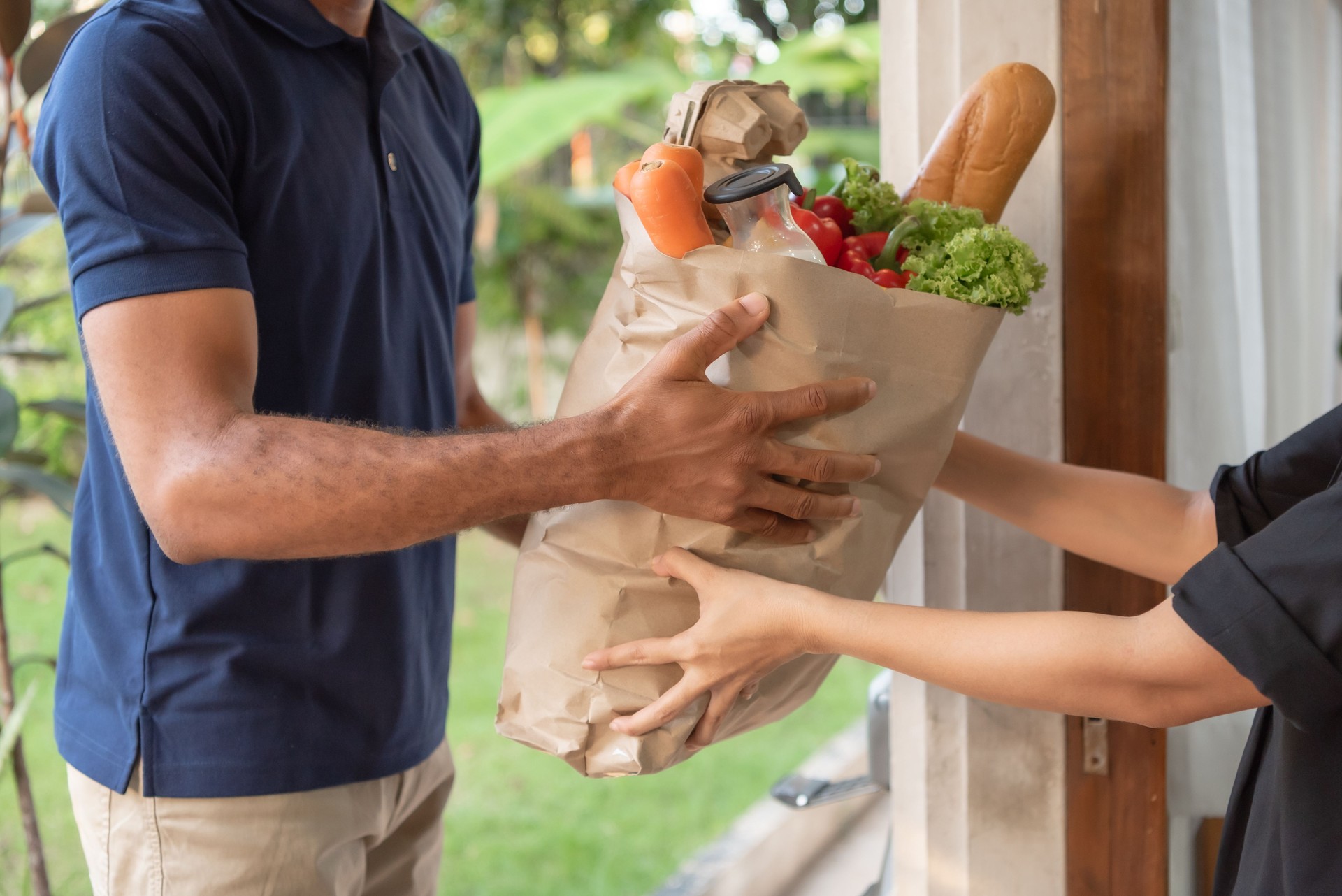 Asian Woman receives a bag full of fresh groceries from African American Delivery man service at a residential doorstep.